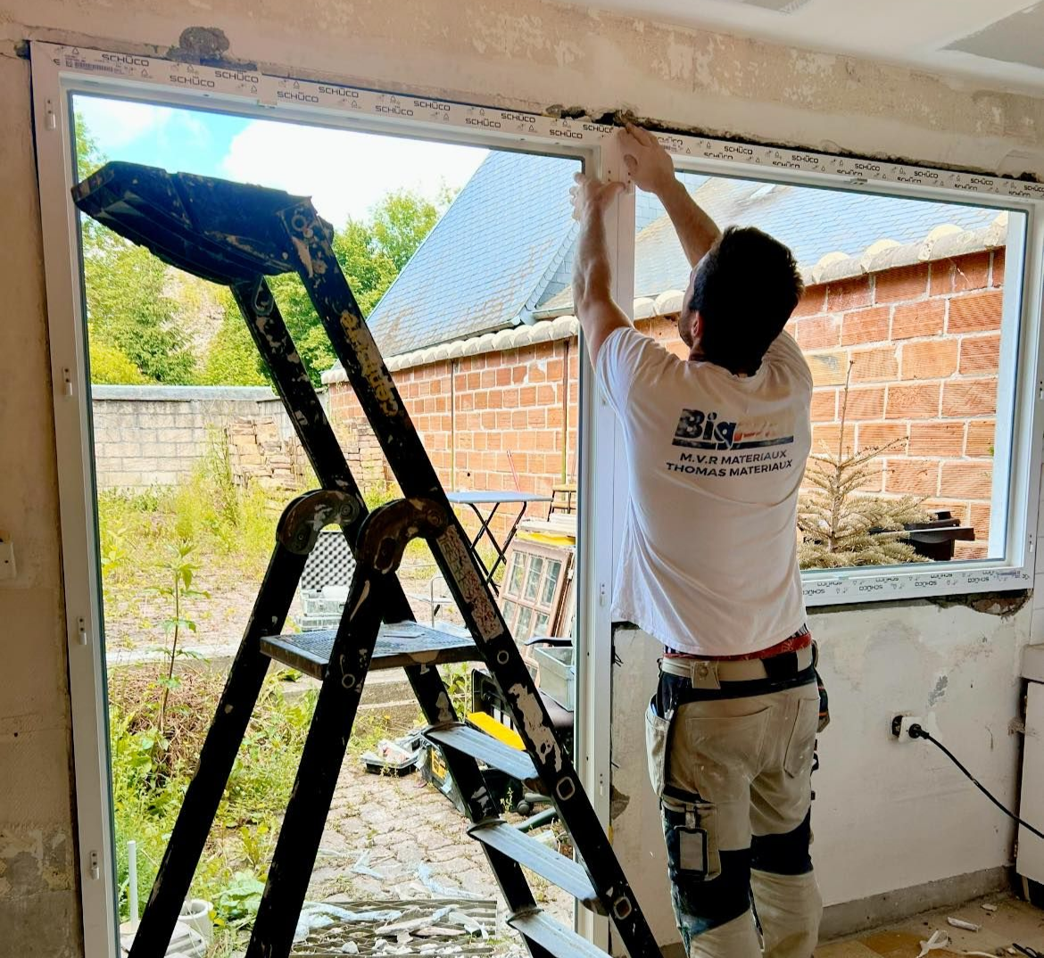 Un homme sur une échelle installe une fenêtre : un mur extérieur en briques est visible en arrière-plan.