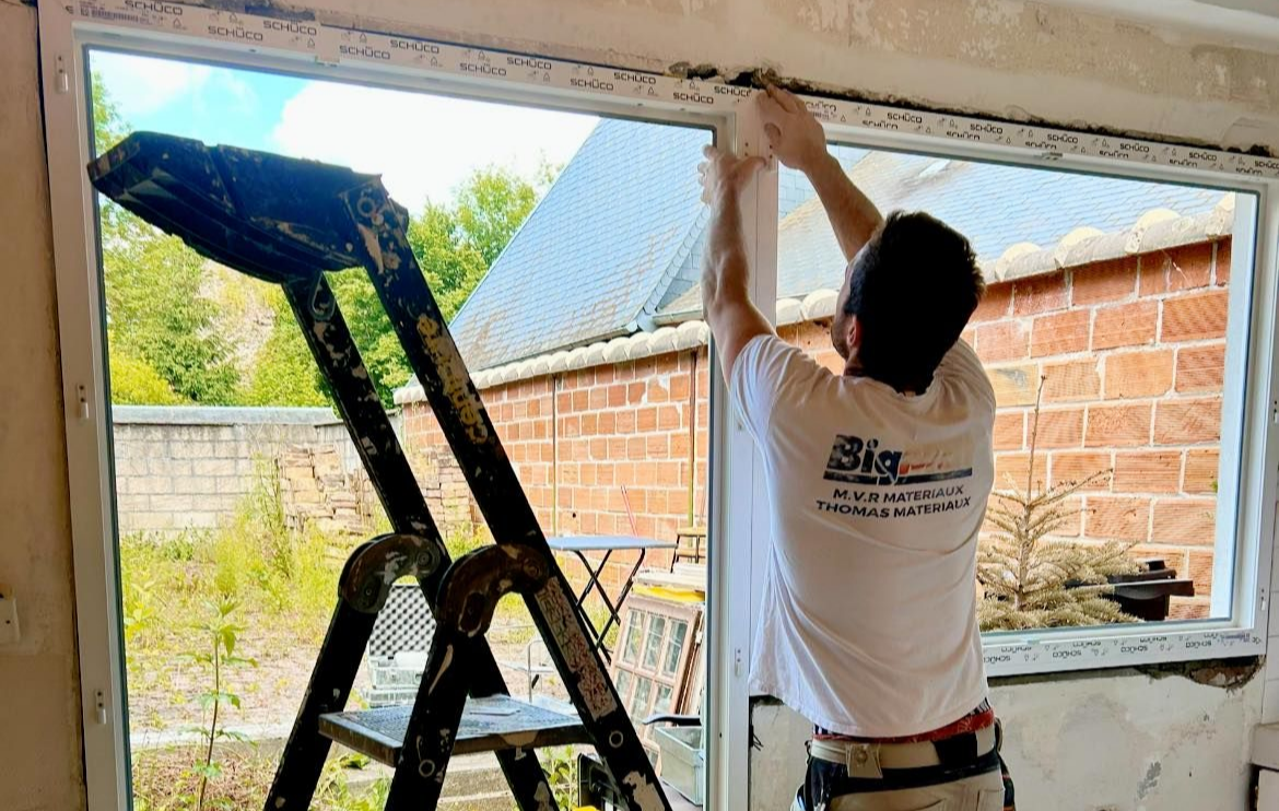 Une personne sur une échelle installe un cadre de fenêtre blanc dans une pièce.