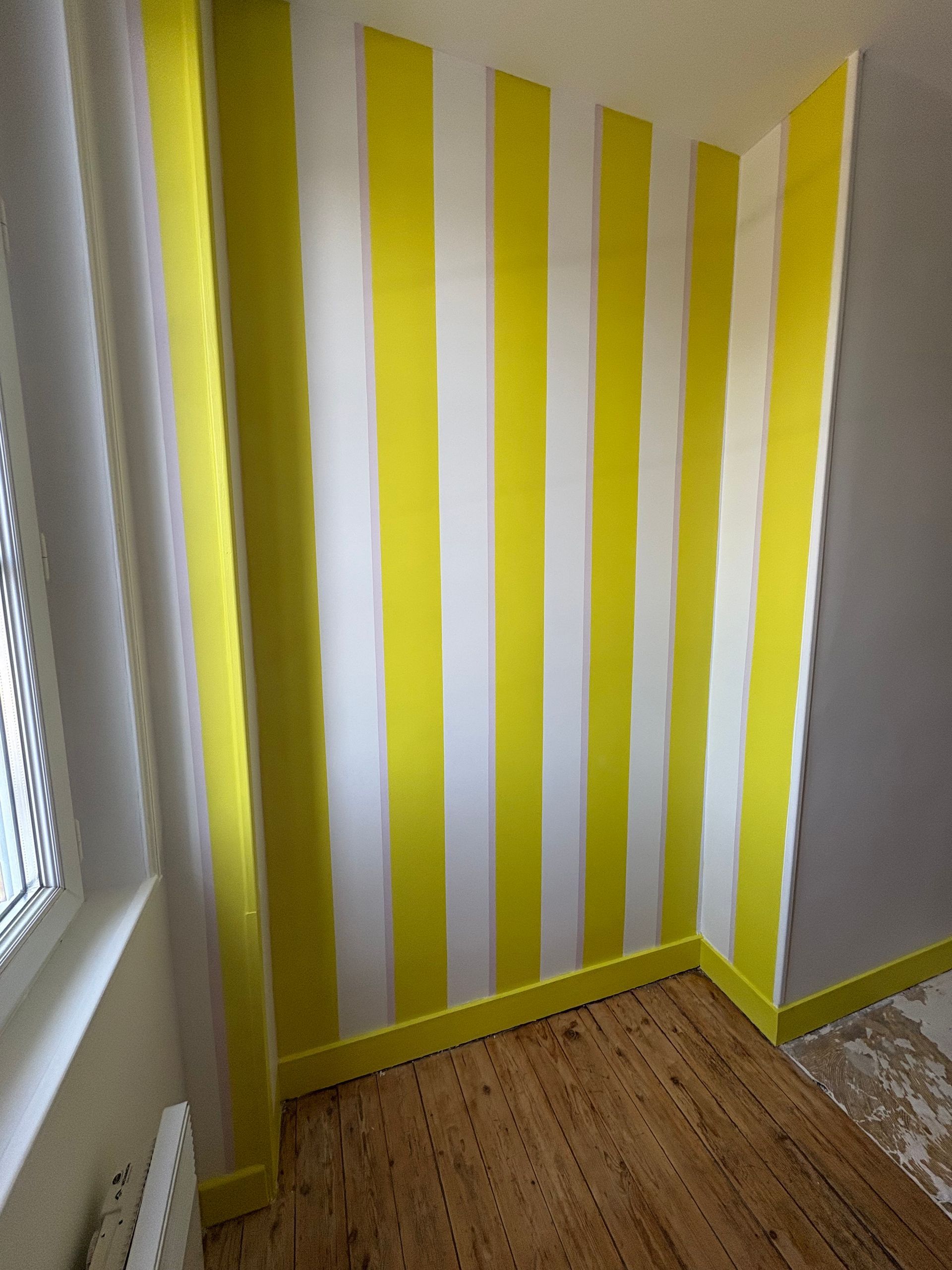 Mur à rayures jaunes et blanches dans une pièce avec parquet et fenêtre.
