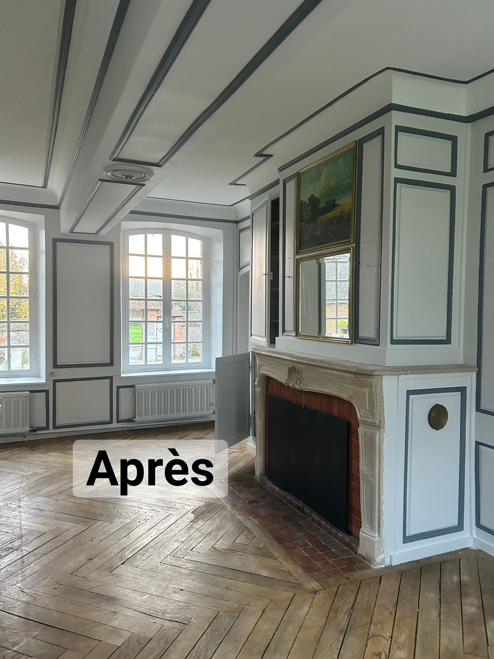 Pièce vide avec parquet, cheminée et fenêtres. Les murs sont blancs avec des moulures bleues.