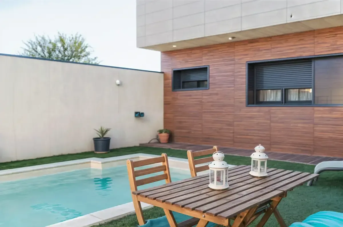 Patio trasero con piscina, mesa y sillas de madera, y faroles decorativos. Pared de ladrillo marrón y paredes beige.