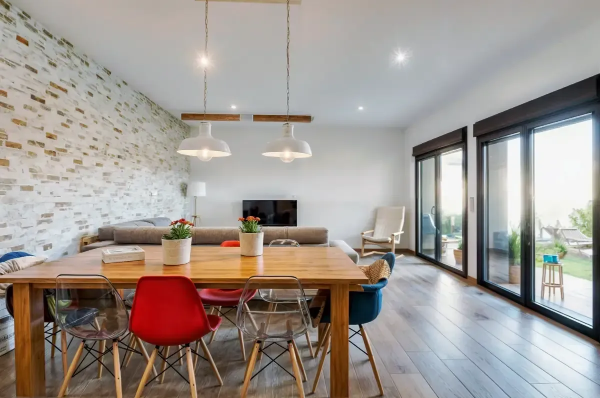 Comedor con mesa de madera, sillas de colores, pared de ladrillos a la vista y puertas corredizas de vidrio.