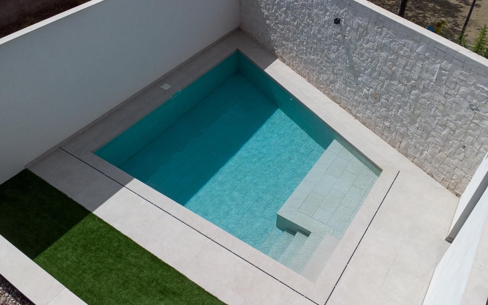 Una pequeña y moderna piscina rectangular con escalones, rodeada de paredes blancas y pavimento de color claro.