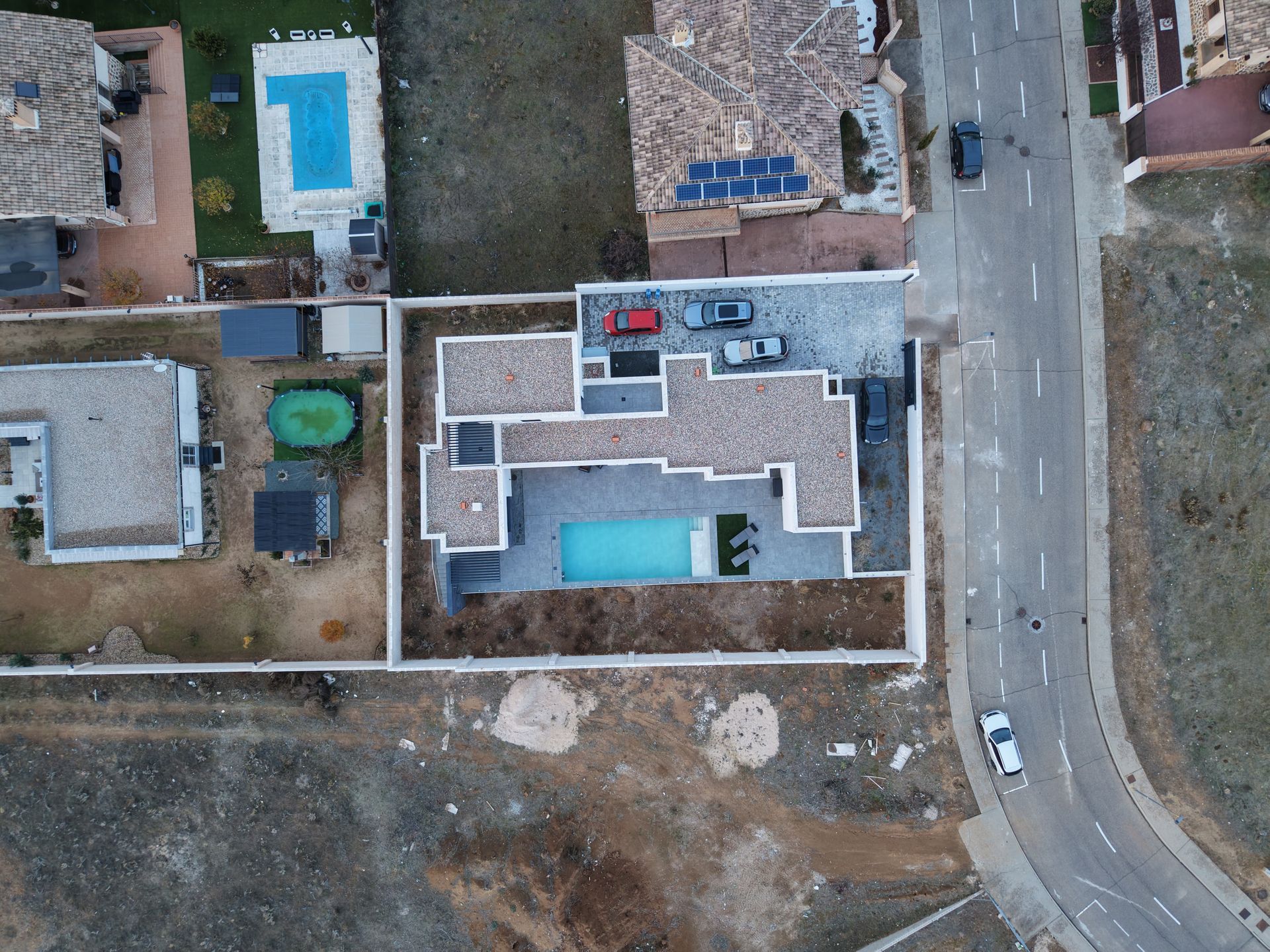 Vista aérea de una casa moderna con piscina, coches aparcados delante y una carretera a la derecha.