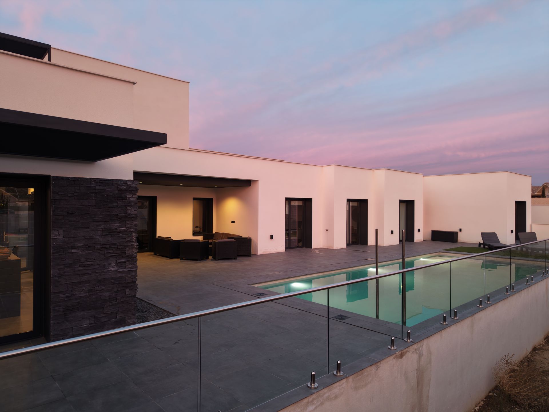 Casa moderna con piscina al anochecer; cielo rosa y azul, muebles de patio oscuros, cerca de piscina de vidrio.