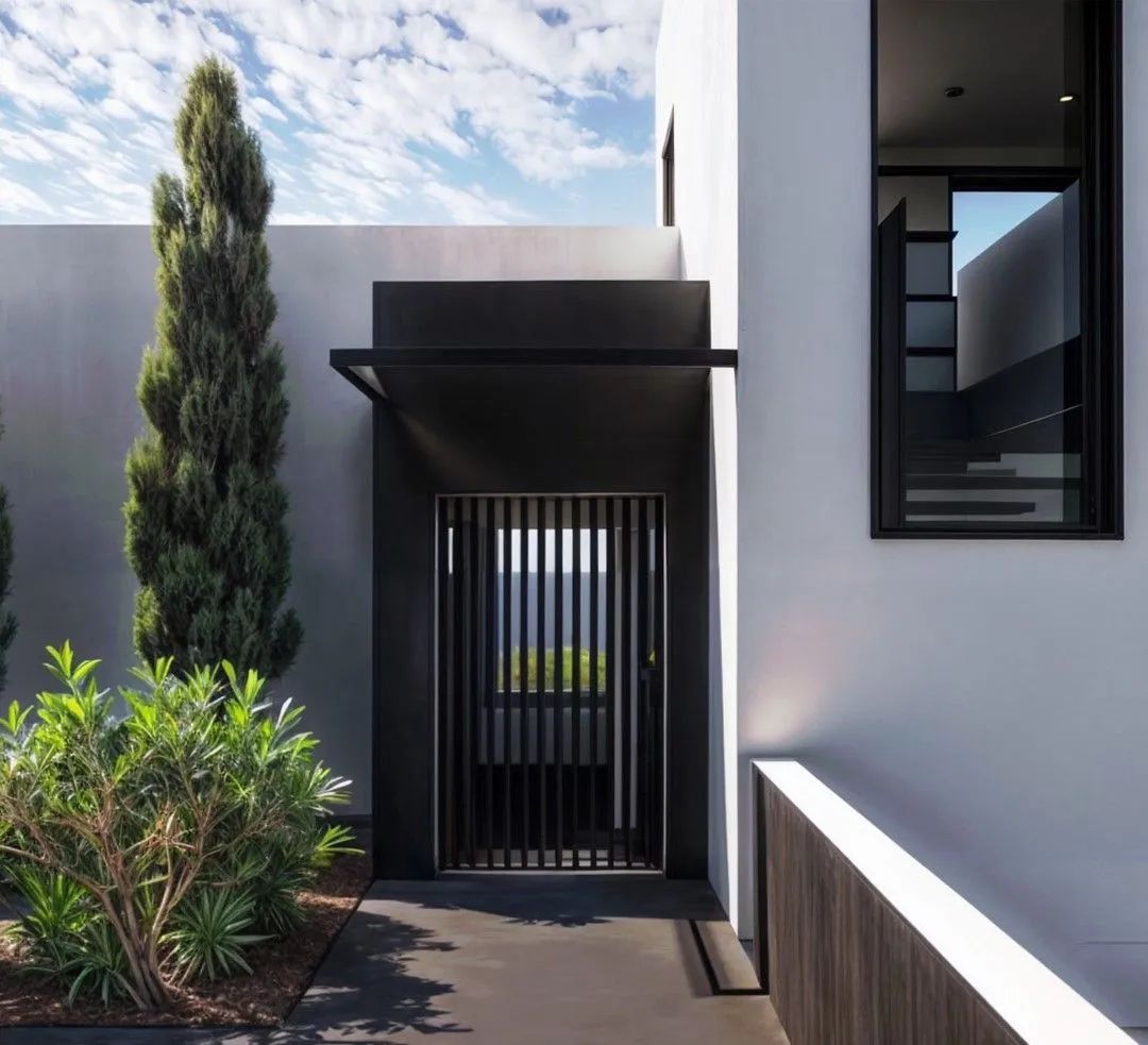 Exterior de casa moderna con entrada oscura, ventana y jardinería contra una pared blanca.