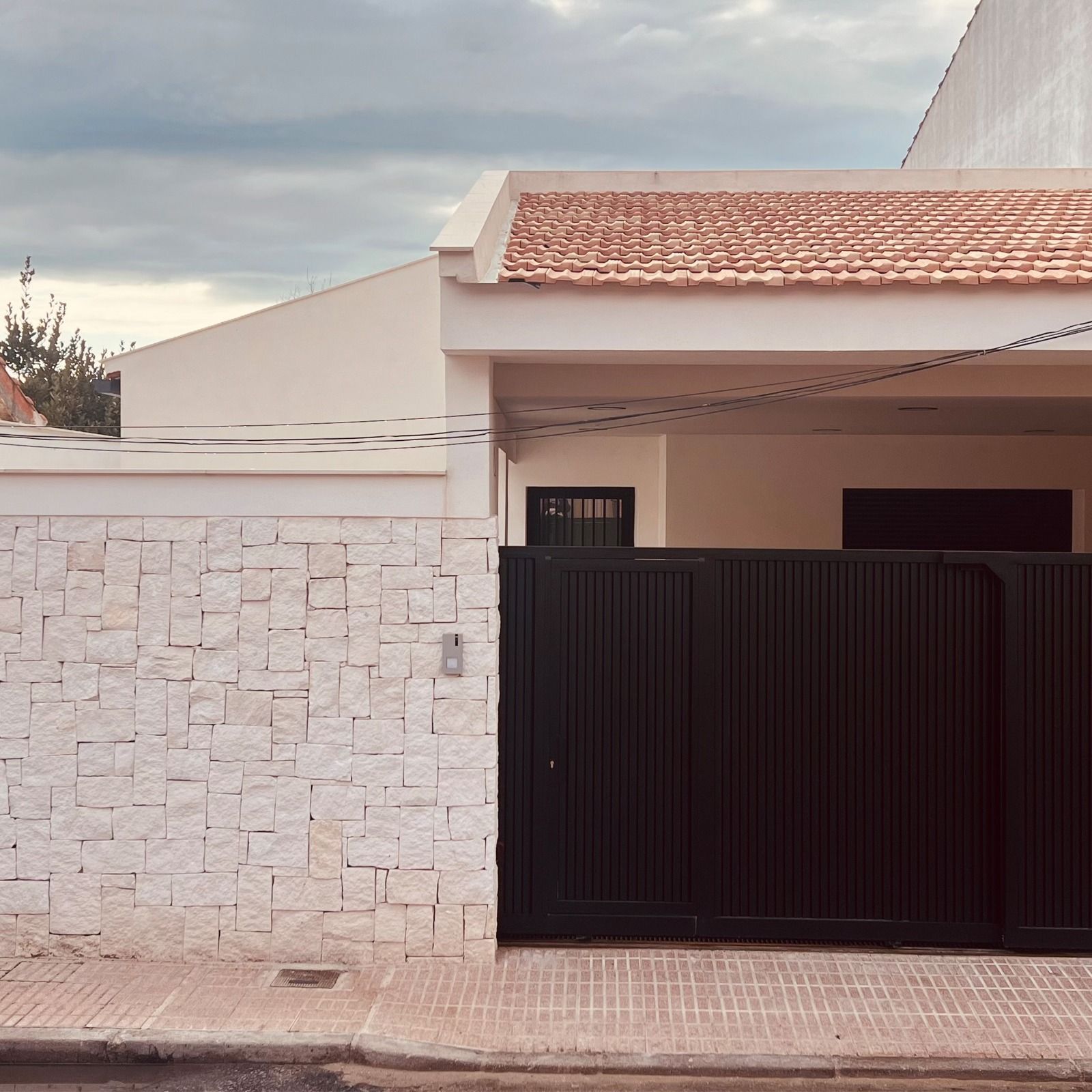 Casa con pared de piedra blanca, portón negro y techo color canela bajo un cielo nublado.
