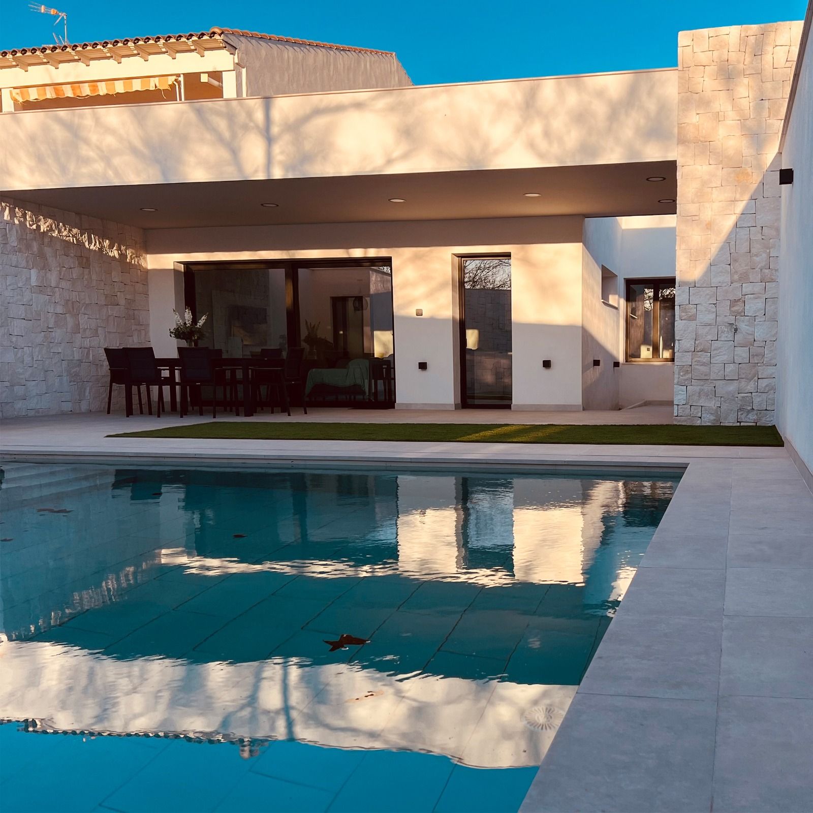 La piscina refleja una casa blanca con patio y muebles oscuros; brilla la luz del sol.