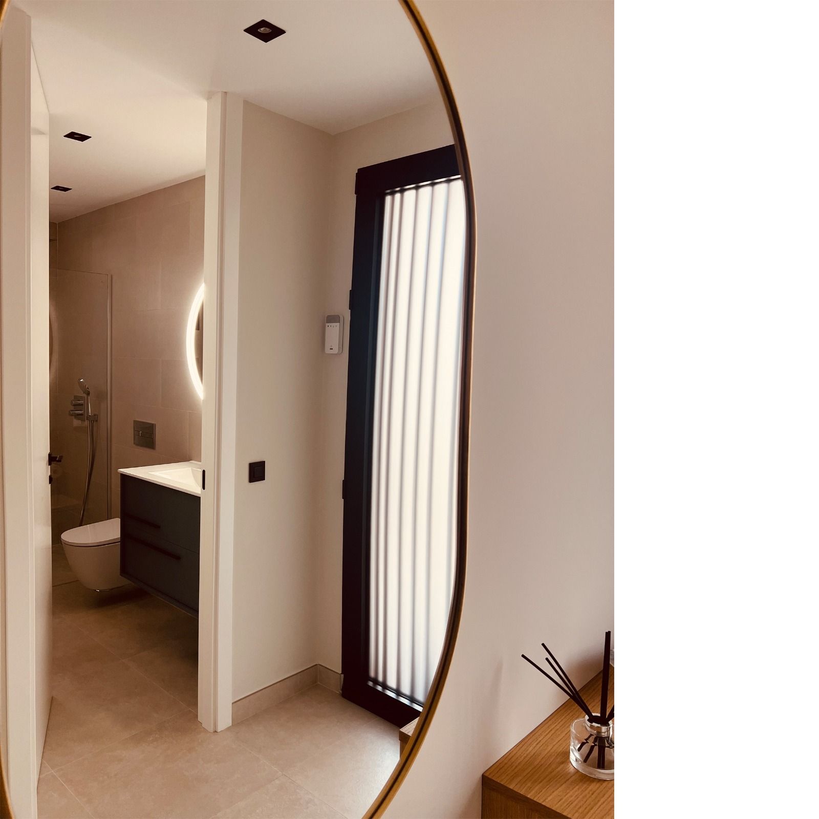 Baño reflejado en un espejo redondo: inodoro, lavabo y ventana en un espacio estrecho.