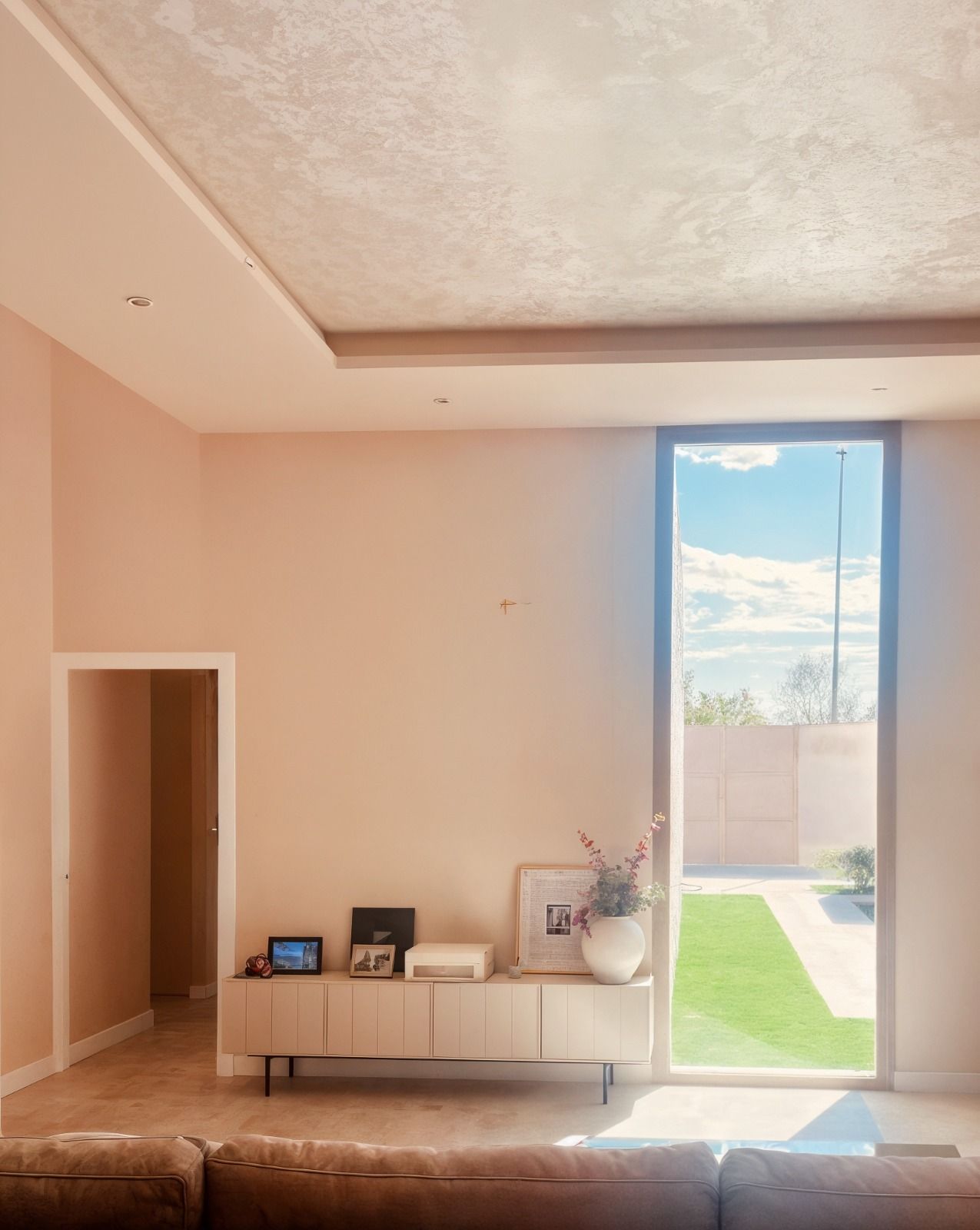 Sala de estar con una ventana alta, paredes de color rosa pálido, un armario de madera clara y vista a un césped.