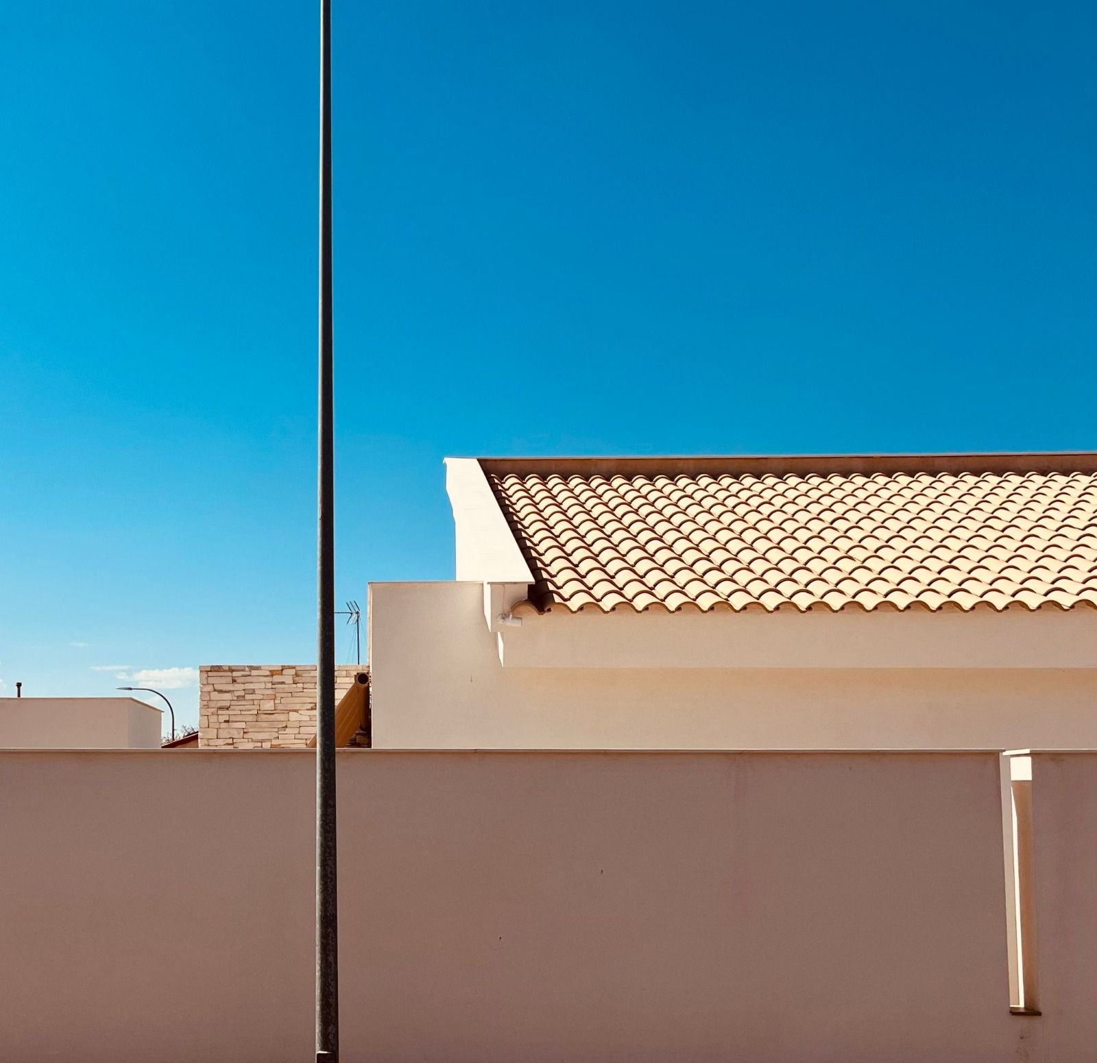 Casa beige con techo de tejas, pared blanca y un poste alto contra un cielo azul vibrante.