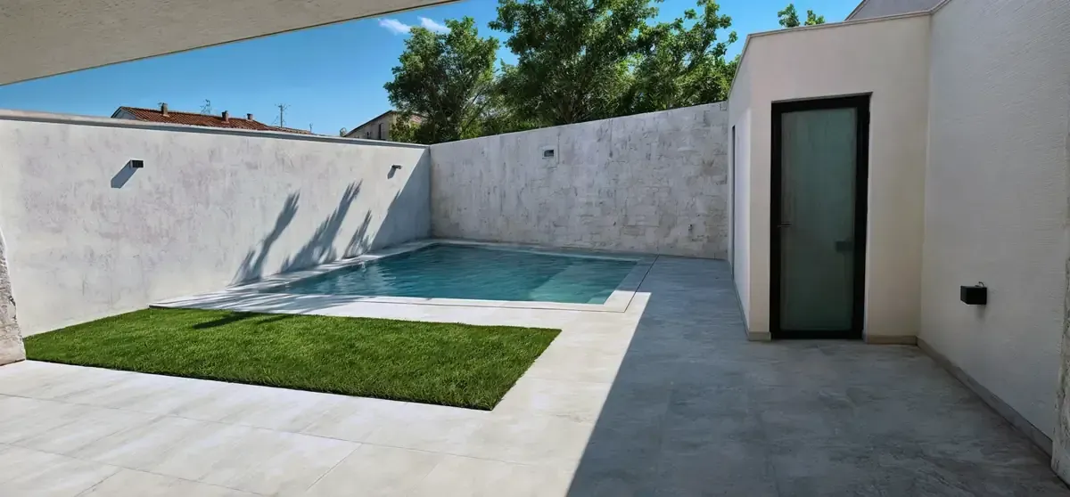 Pequeña piscina en un patio con zona de césped y puerta. Paredes blancas y un día soleado.
