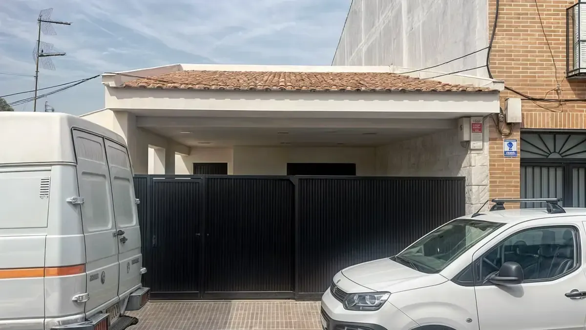 Casa blanca con portón negro y vehículos estacionados afuera.