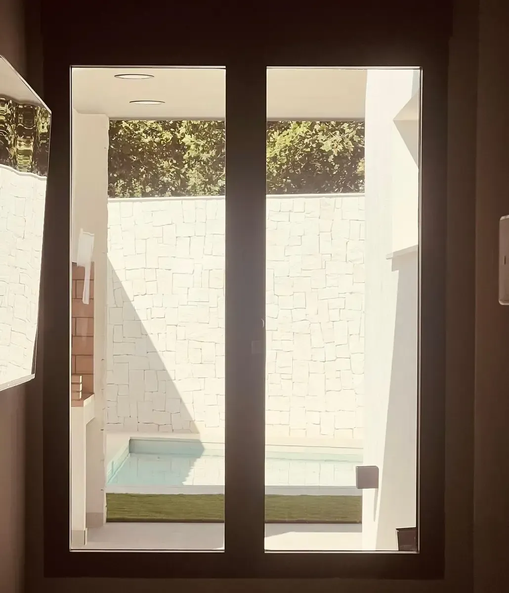 Vista a través de una ventana: pequeña piscina rodeada de paredes blancas, con vegetación encima.