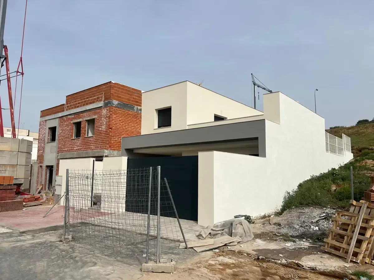 Sitio de construcción: casa moderna con exterior gris y blanco, sección de ladrillo sin terminar, puerta negra, cerca y cielo azul.