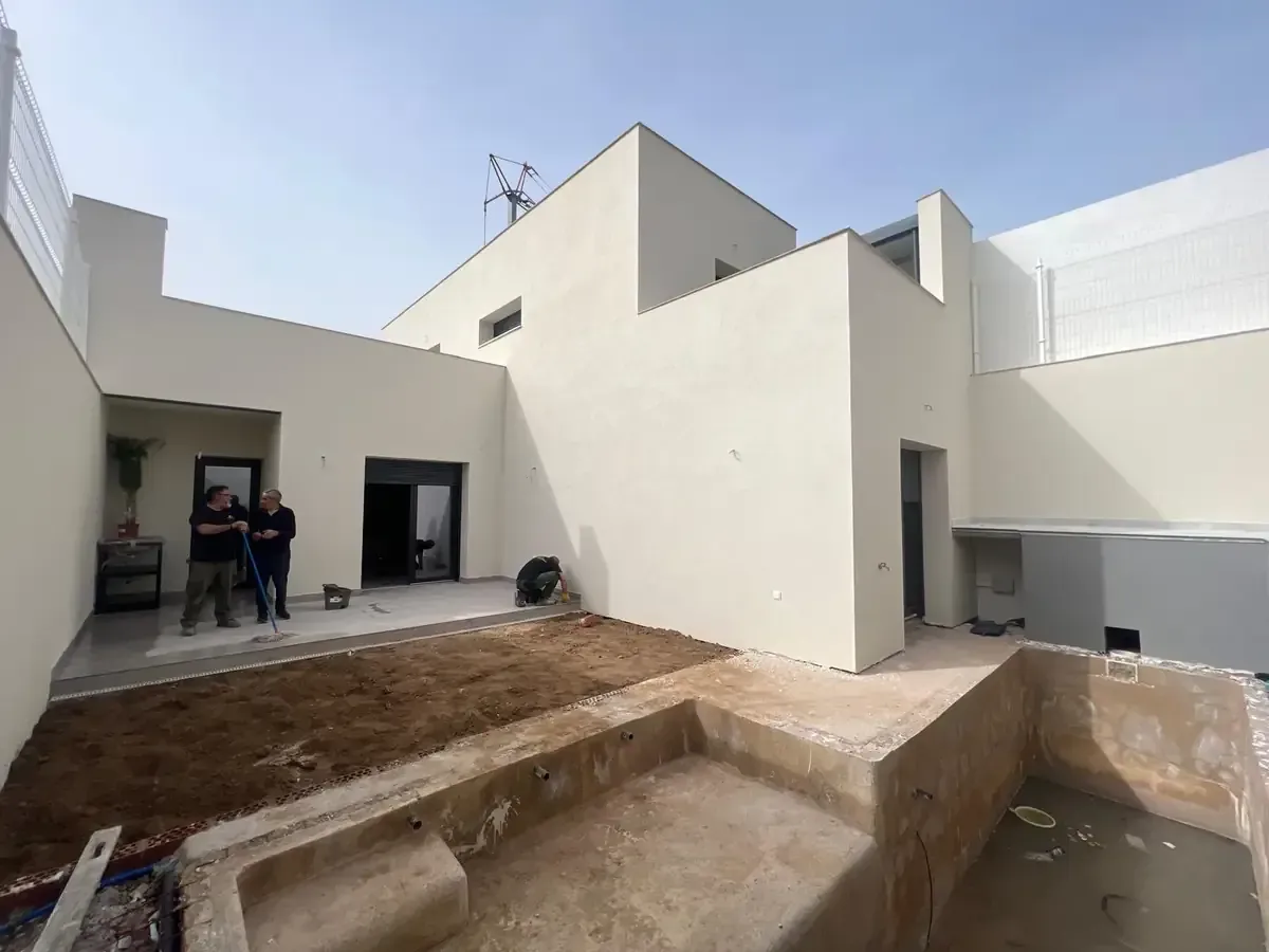 Vista exterior de una casa moderna en construcción con una piscina sin terminar y dos personas paradas afuera.