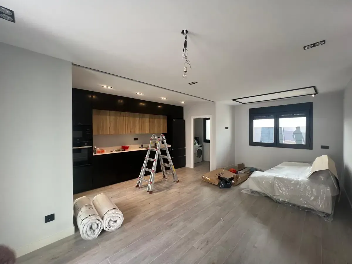 Apartamento moderno en reforma: cocina con muebles negros, detalles en madera, escalera y cama revestida de plástico.