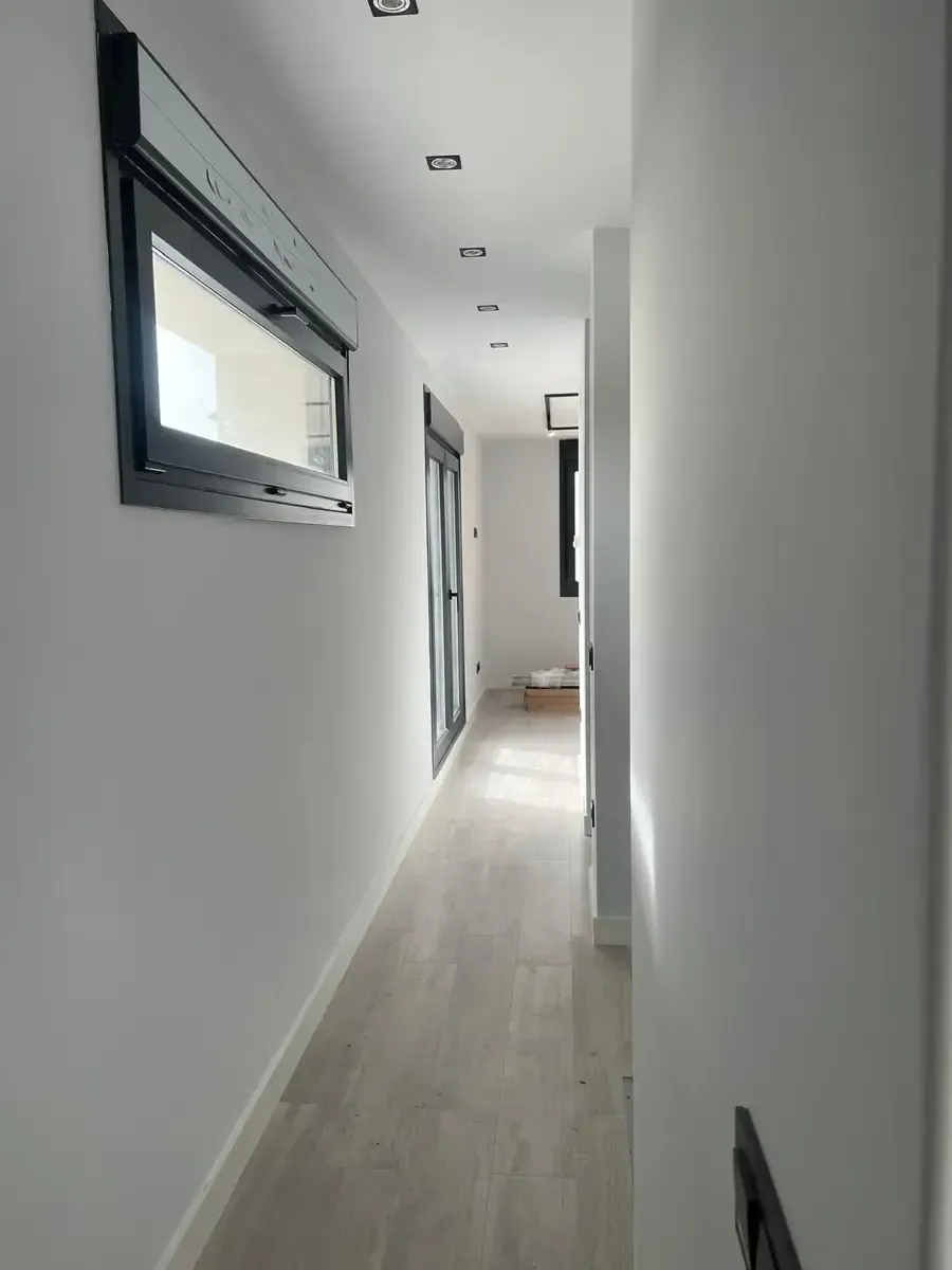 Pasillo estrecho con paredes blancas, suelo gris e iluminación empotrada, con ventanas con marcos negros.