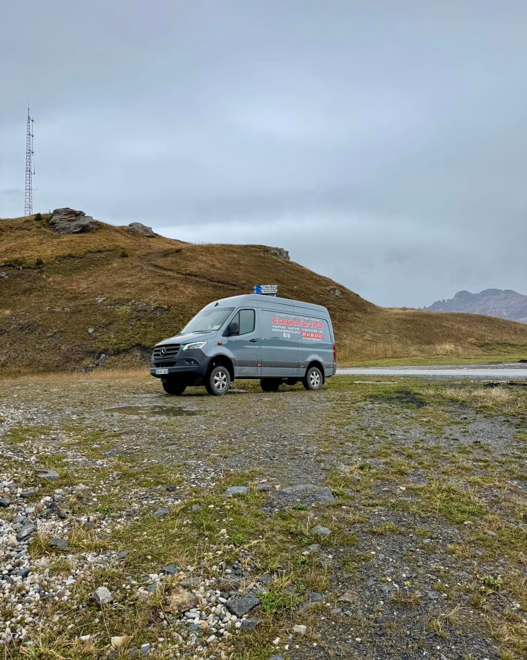 Camionnette grise garée sur du gravier, paysage montagneux avec ciel couvert.