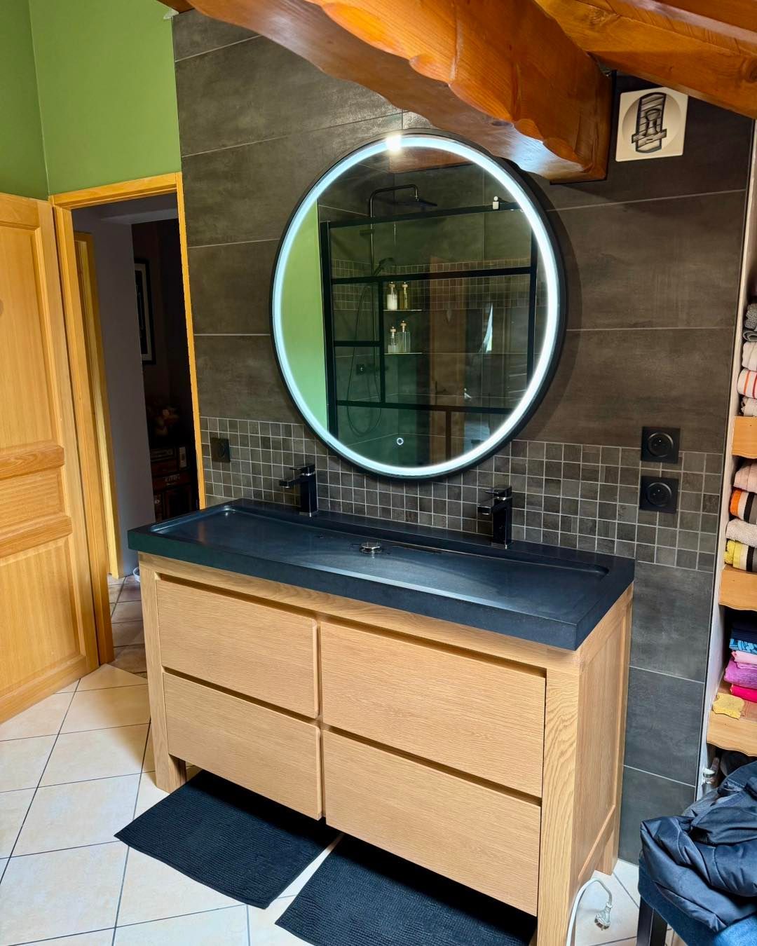 Salle de bain avec vanité en bois, comptoir noir, miroir rond éclairé et dosseret en carrelage foncé.