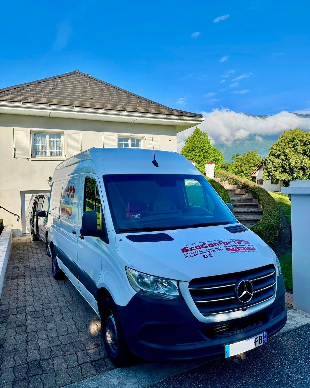 Fourgonnette Mercedes blanche garée dans l'allée devant une maison, vue sur la montagne en arrière-plan.