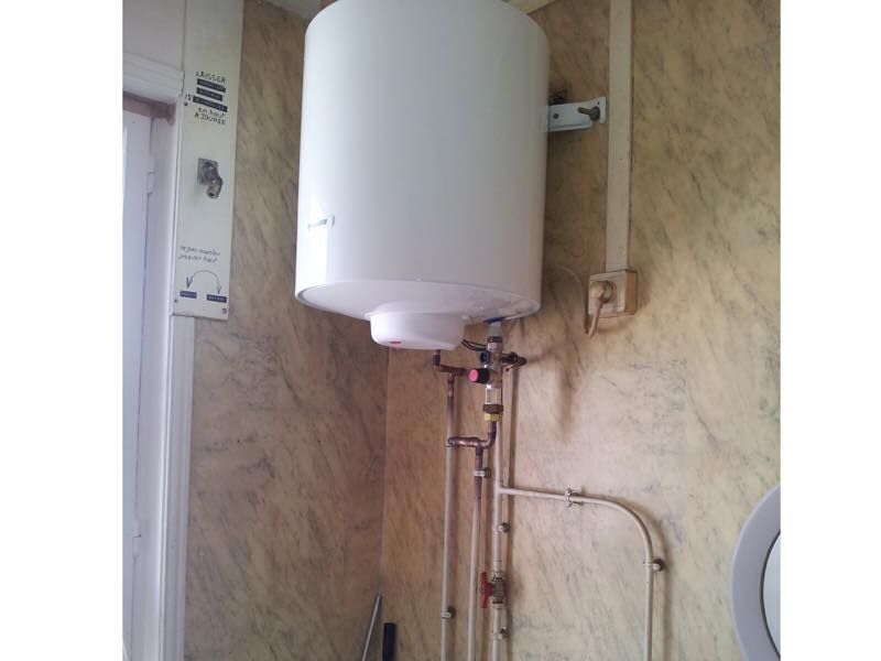 Chauffe-eau blanc fixé au mur, avec tuyaux et vannes en dessous. Murs beiges.