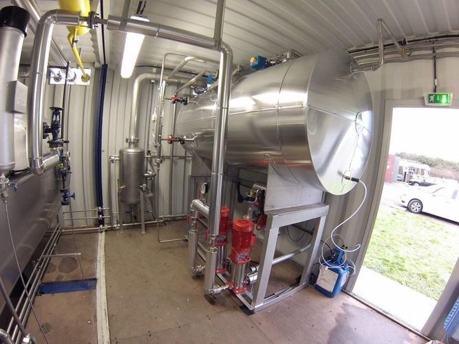 Équipement industriel en acier inoxydable dans une petite pièce blanche. On y voit des tuyaux, des réservoirs et une pompe rouge.