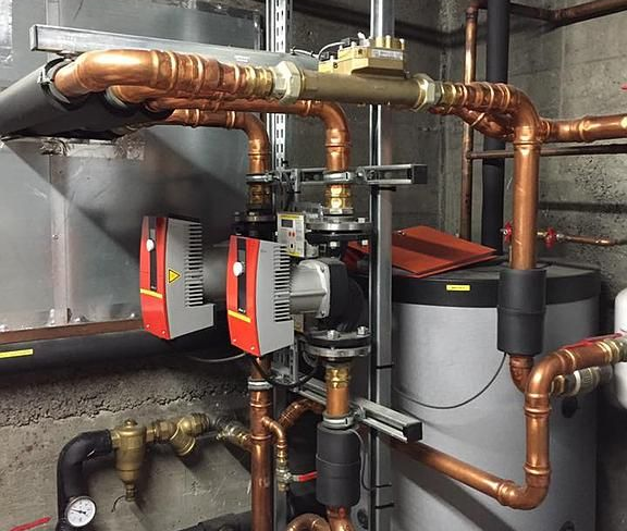 Tuyaux et pompes en cuivre dans une salle mécanique avec réservoirs gris, mur de briques brunes et tuyaux isolés.