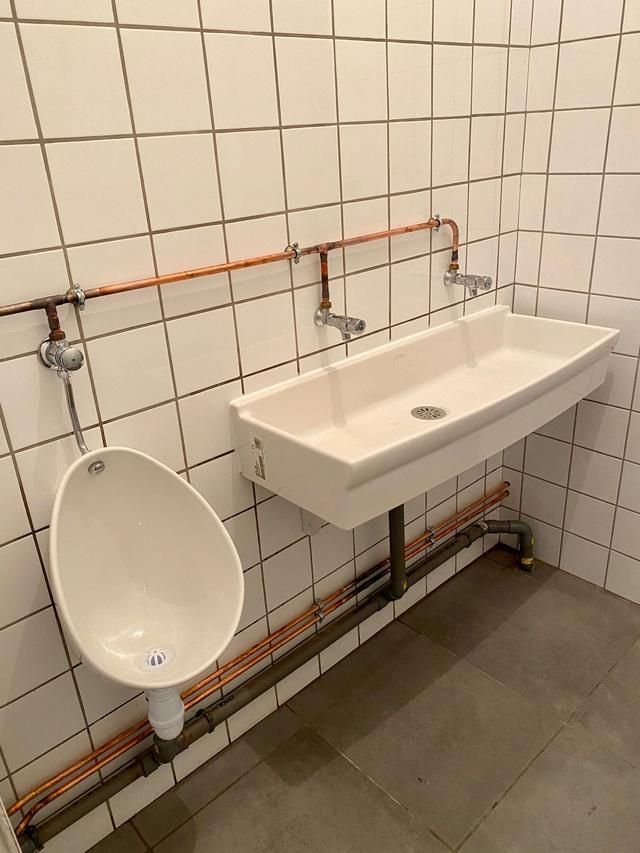 Salle de bain avec urinoir, lavabo et plomberie en cuivre apparente. Murs carrelés blancs et sol gris.