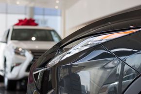 Primer plano del faro de un coche gris oscuro, otro SUV blanco con un lazo rojo al fondo, en una sala de exposición de automóviles.