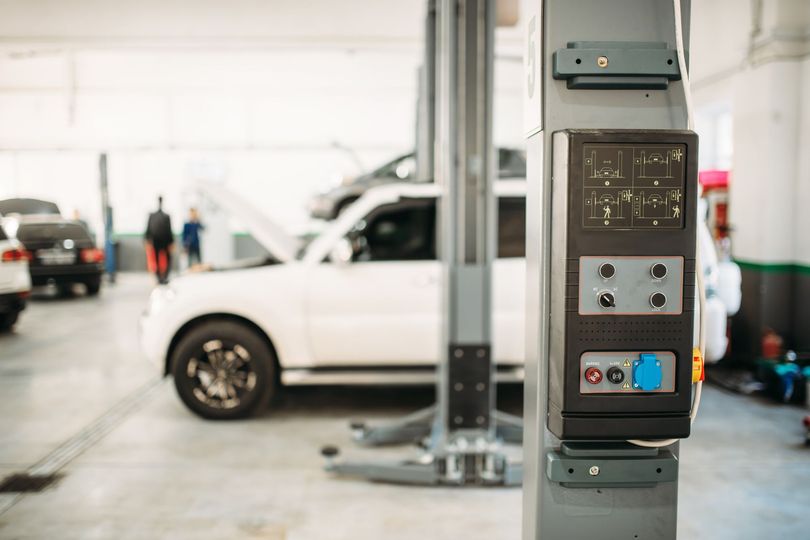 Panel de control de un elevador de coches en un garaje, con un todoterreno blanco y otros vehículos al fondo.