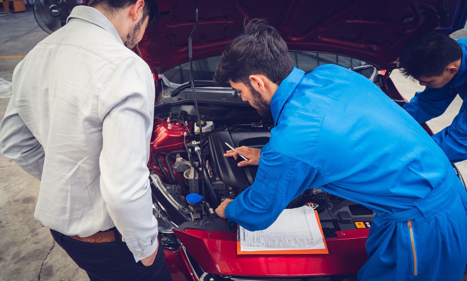 Dos mecánicos con monos azules y un cliente con chaqueta blanca inspeccionan el motor de un coche rojo.