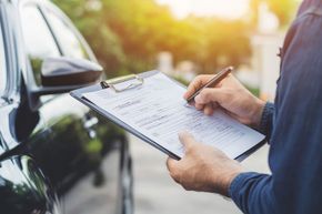 Dos mecánicos con monos azules y un cliente con chaqueta blanca inspeccionan el motor de un coche rojo.