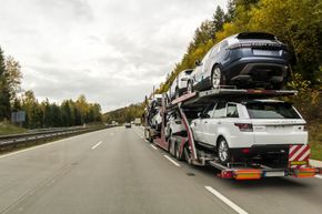 Transportador de automóviles que transporta vehículos todoterreno en una carretera con árboles al fondo bajo un cielo nublado.