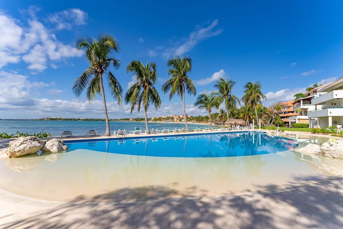 Tropical resort pool with palm trees, sandy beach, blue ocean, and bright sunny sky