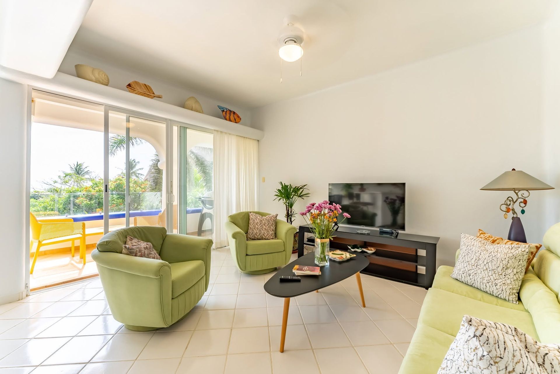 Bright living room with green chairs, sofa, TV, coffee table, and sliding glass doors to a sunny patio.