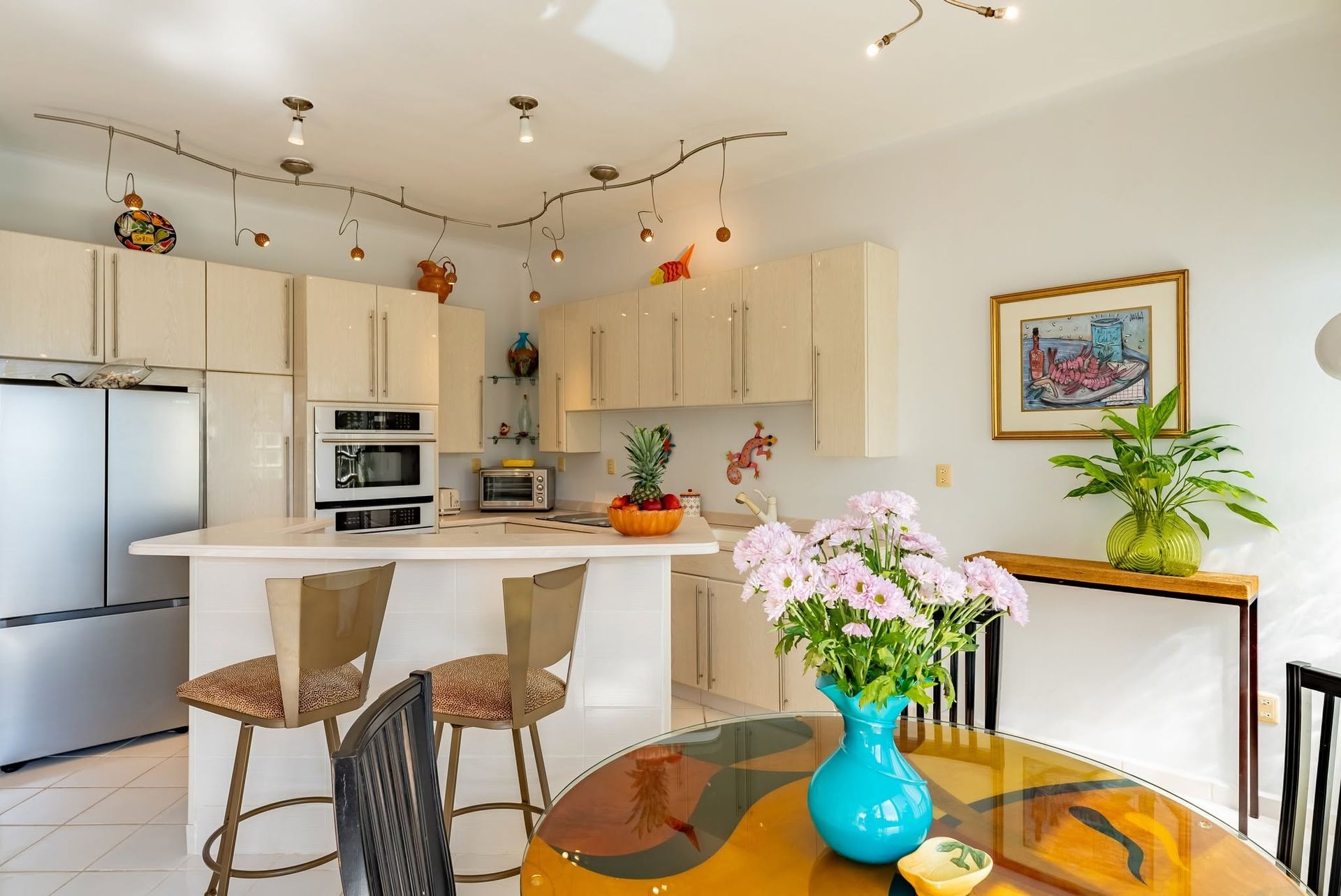 Bright kitchen and dining area with white cabinets, wicker stools, flowers, and a glass table