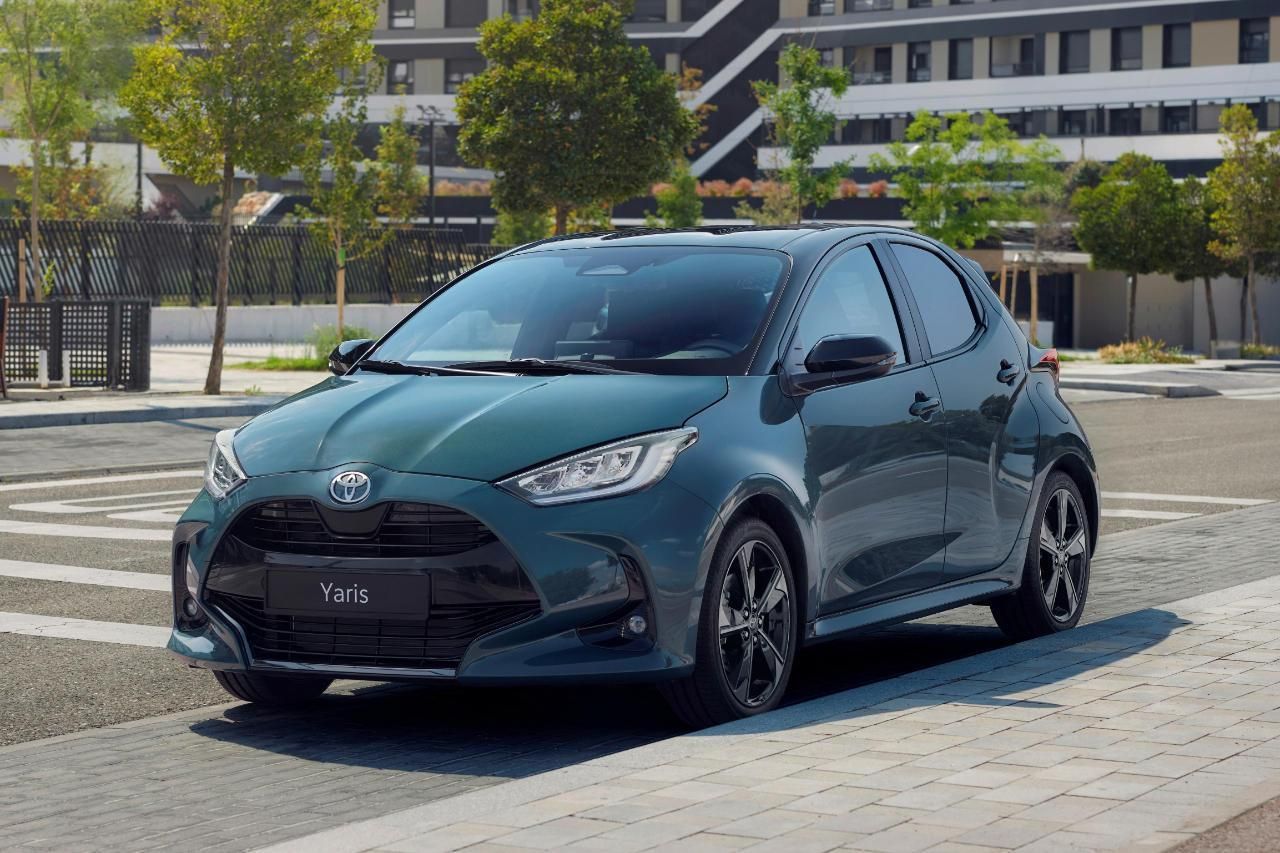 Une Toyota Yaris bleu sarcelle foncé est garée sur une aire pavée, avec des immeubles d'appartements en arrière-plan.