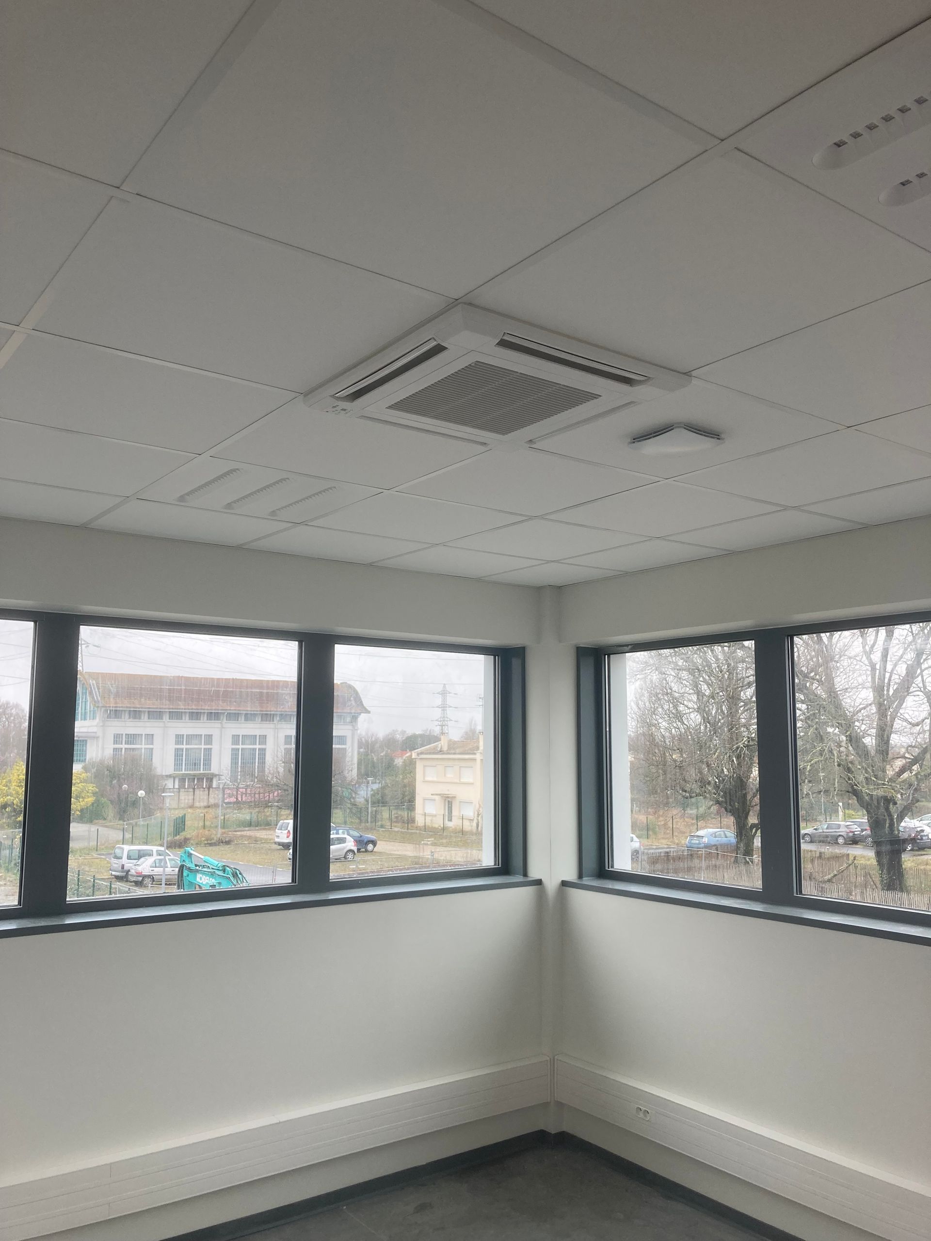 Coin bureau intérieur avec grille d'aération au plafond, grandes fenêtres et vue sur l'extérieur.