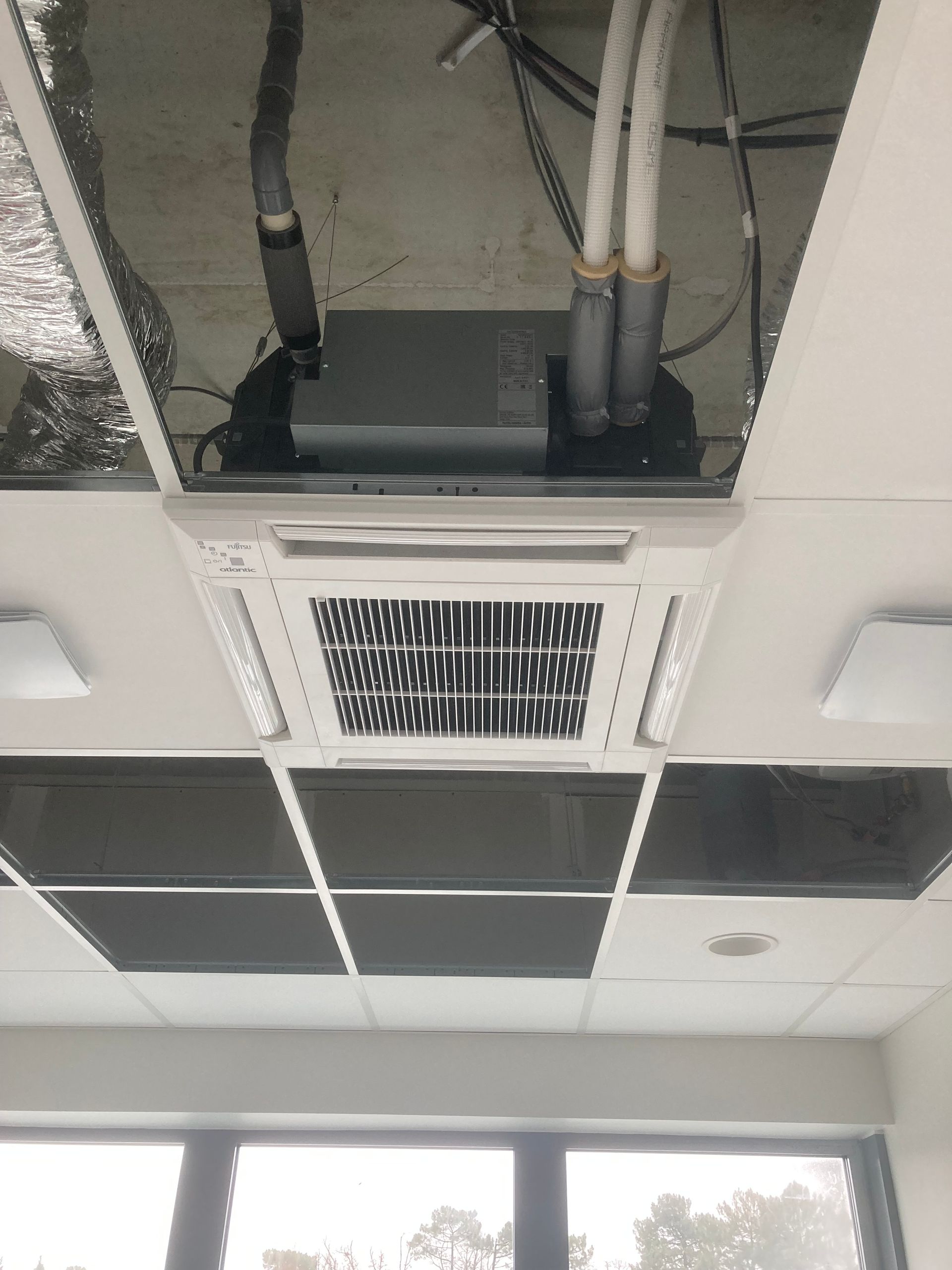 Unité de climatisation fixée au plafond dans un faux plafond, avec conduits et luminaires visibles.