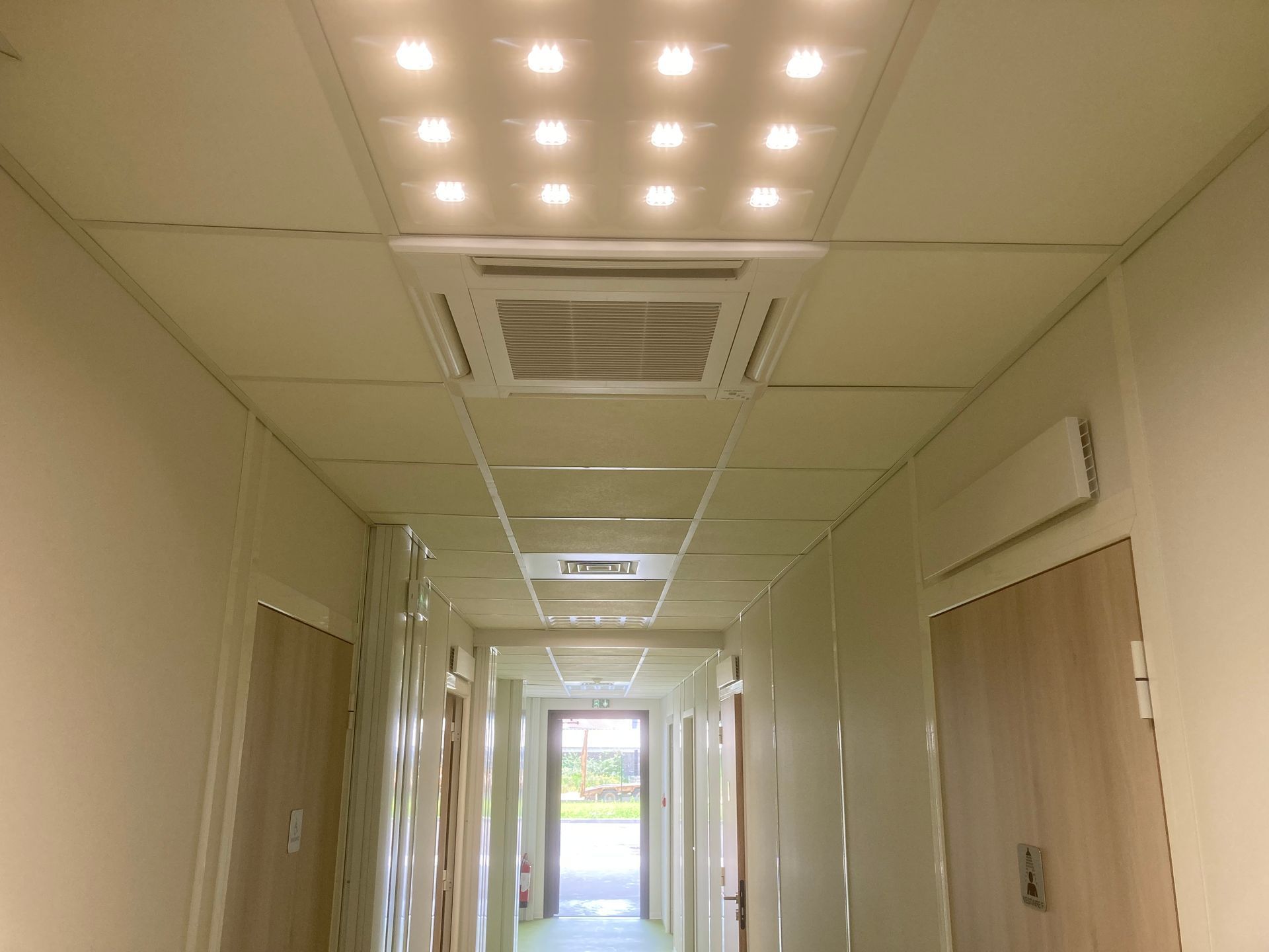 Couloir avec plafond en dalles blanches, unité de climatisation, spots encastrés et portes.