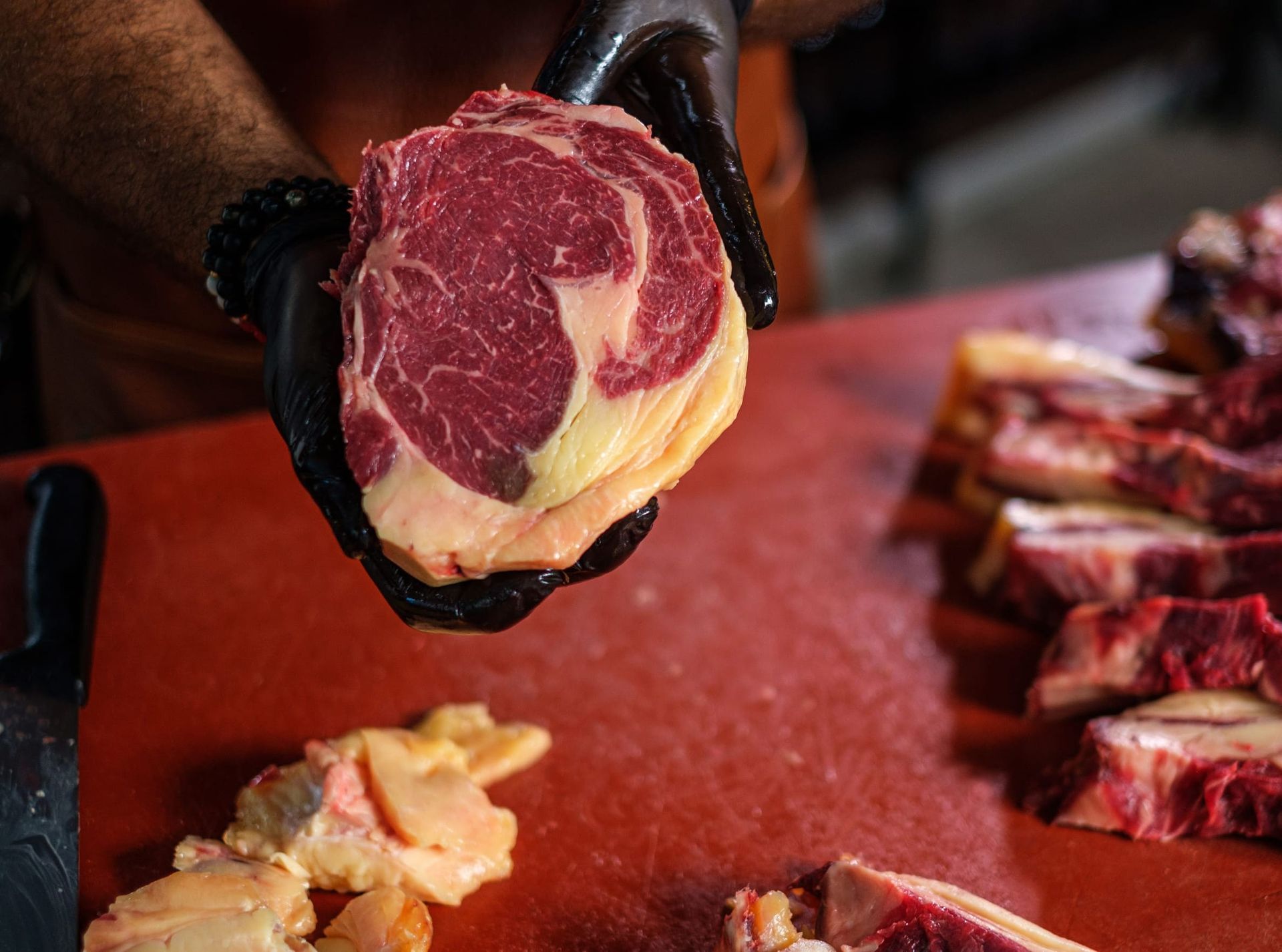 Unas manos enguantadas sostienen un filete crudo sobre una tabla de cortar de carnicero, con carne en lonchas cerca.