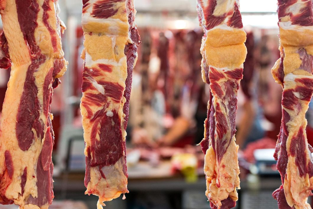 Tiras de carne cruda colgando de un puesto de mercado, con compradores borrosos al fondo.