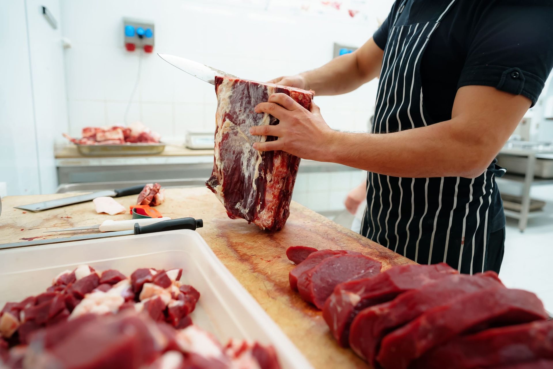 Un carnicero recorta un gran trozo de carne cruda sobre una mesa de trabajo en una carnicería luminosa.