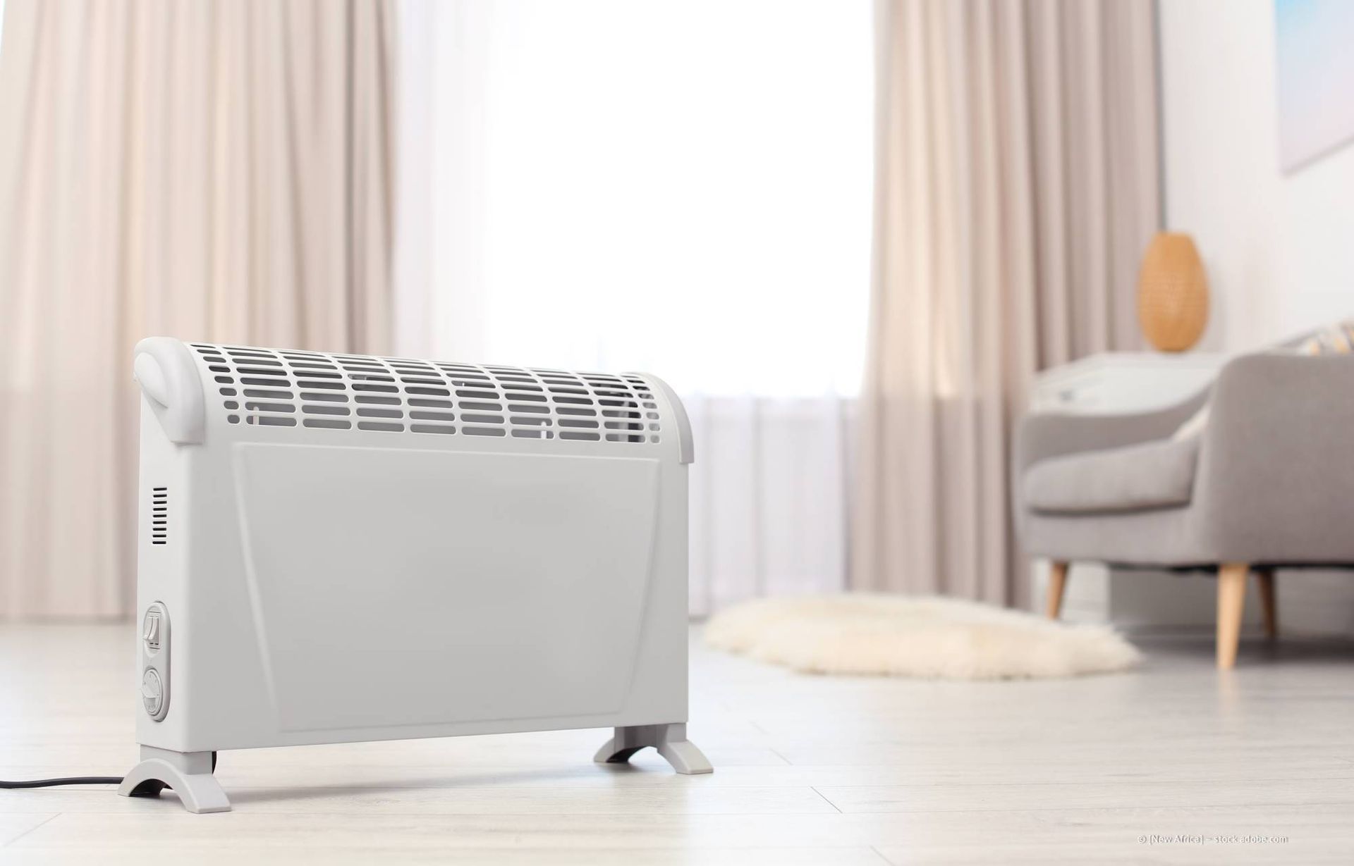 Weißer elektrischer Heizstrahler auf hellem Holzboden in einem Wohnzimmer mit Fenster und Couch.