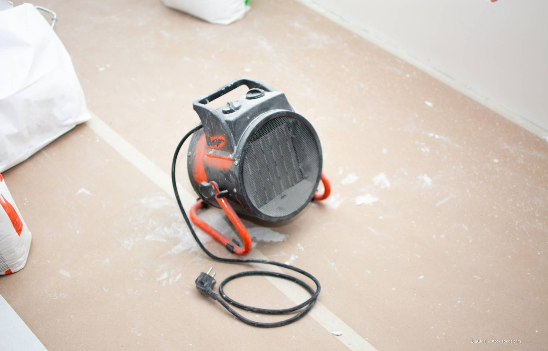 Elektrischer Ventilator auf einem mit weißem Staub bedeckten Boden; orangefarbene Griffe, schwarzes Kabel.