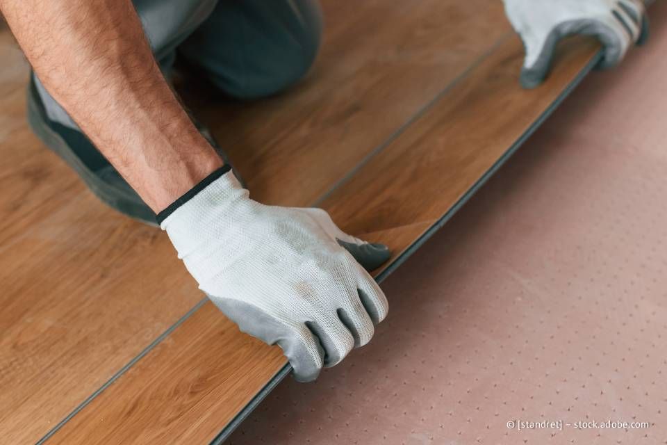 Person mit Handschuhen beim Verlegen eines Bodenbelags in Holzoptik.