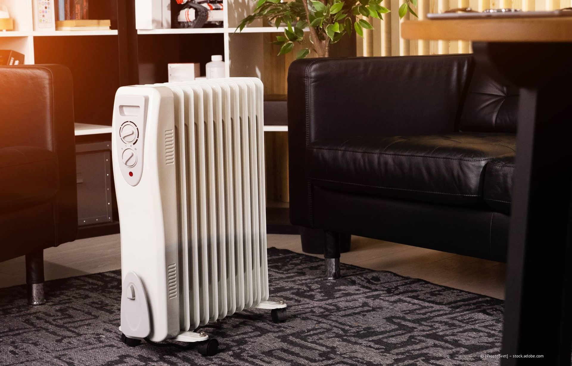 Weißer, mit Öl gefüllter Heizstrahler auf einem Teppich in einem Wohnzimmer, neben schwarzen Ledermöbeln.