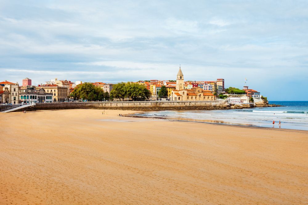 Una vasta playa de arena conduce a un pueblo costero con una prominente torre de iglesia bajo un cielo suave y nublado.