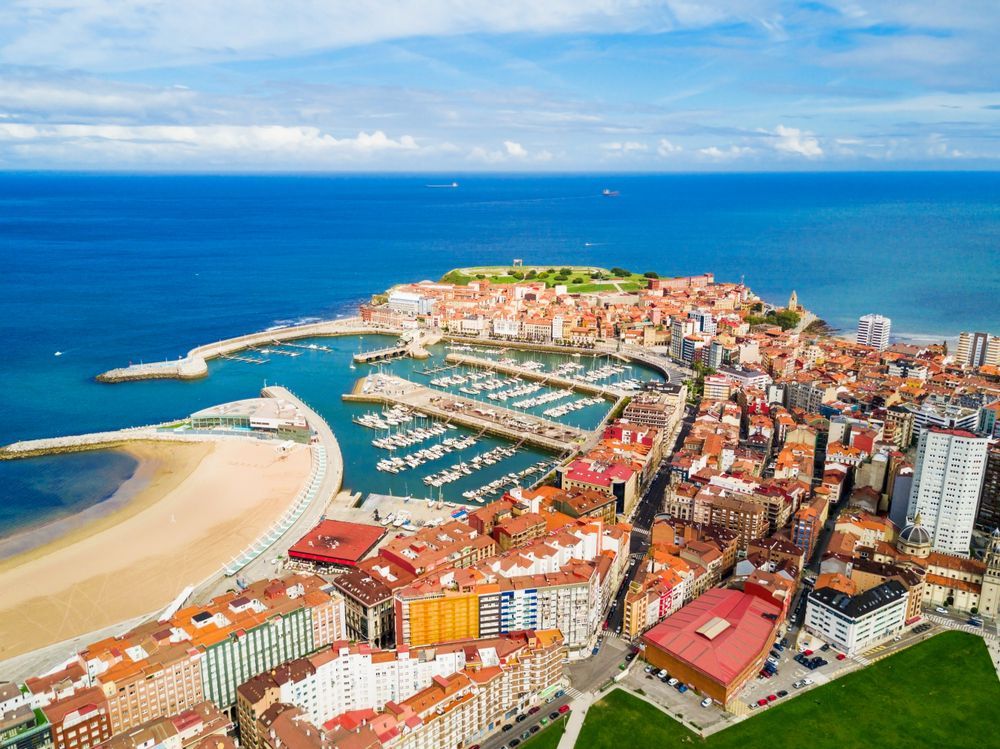 Vista aérea de la ciudad costera de Gijón, España, donde se aprecian su puerto, barcos, playa de arena y edificios urbanos circundantes.